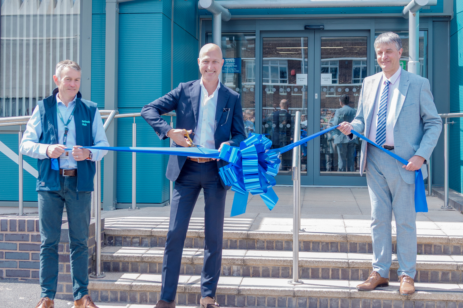 A group of individuals are cutting a blue ribbon in front of a building entrance, celebrating an event.