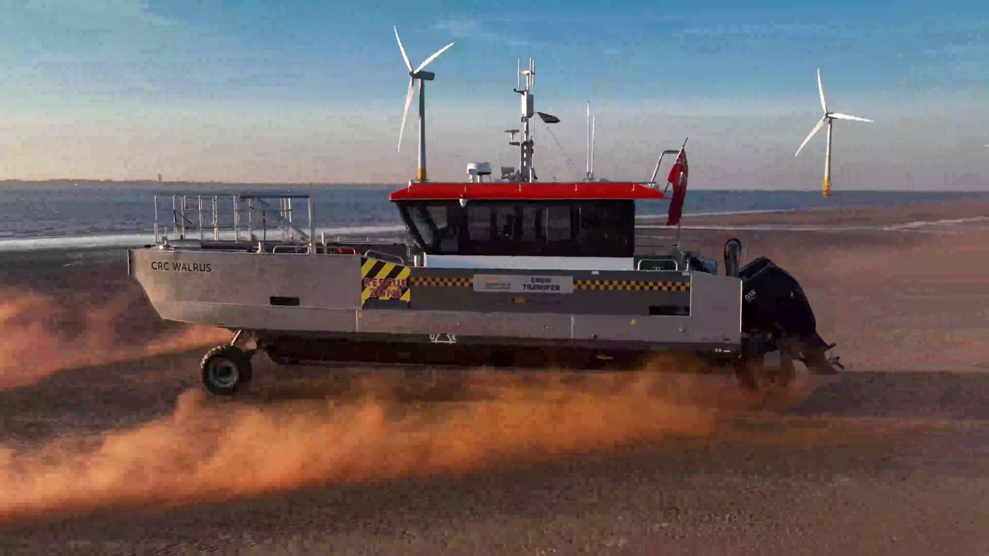 A boat named 'CRC Walrus' on sandy terrain with wind turbines and water in the background.