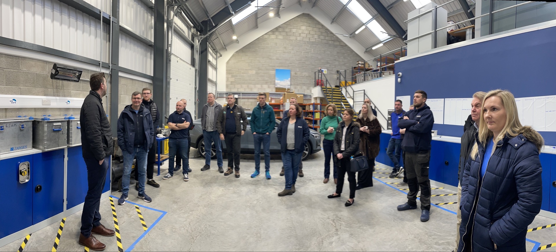 A group of people gathered in a bright industrial space, engaged in discussion near workstations and storage units.