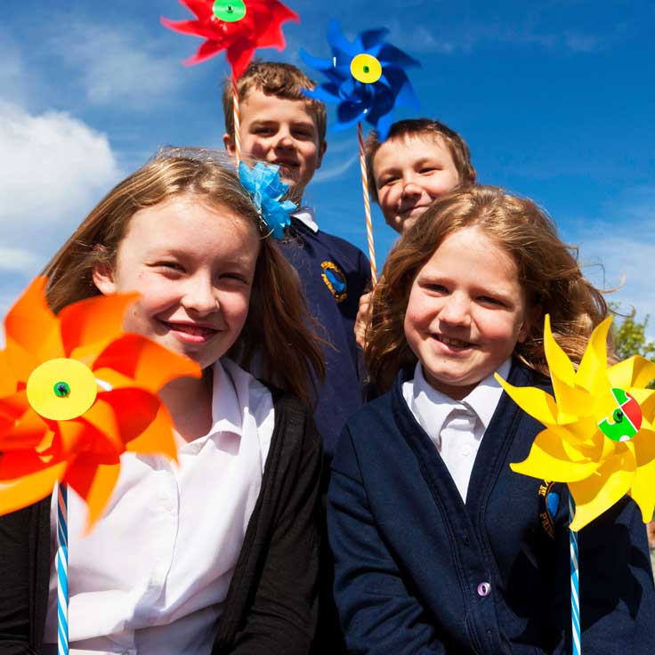 A group of children holding brightly coloured pinwheels against a clear blue sky, showcasing a fun outdoor activity.