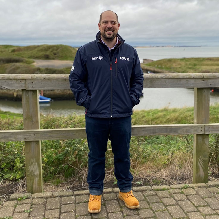 A person stands by a wooden fence near a waterway, wearing a navy RWE jacket, jeans, and tan boots. Grass-covered hills are in the background. Portrait of Jonny Brownsteen.