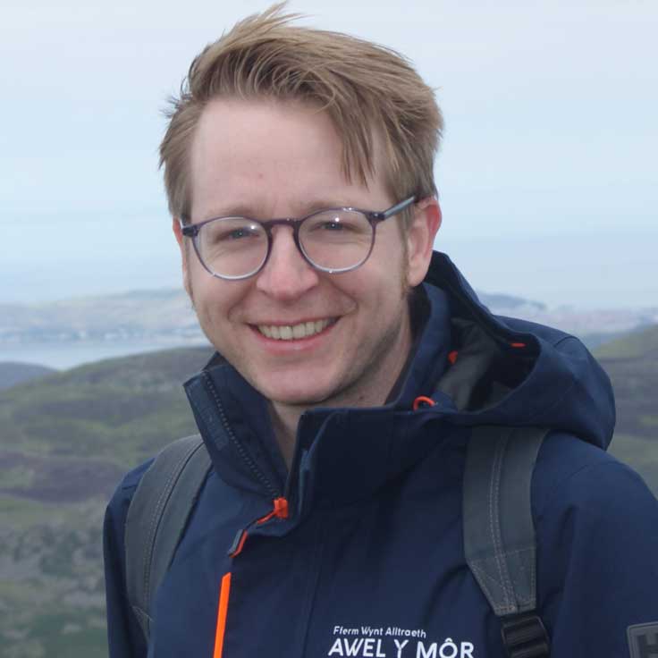 A person stands outdoors, wearing a dark jacket with an orange zipper, against a scenic backdrop of hills and water.