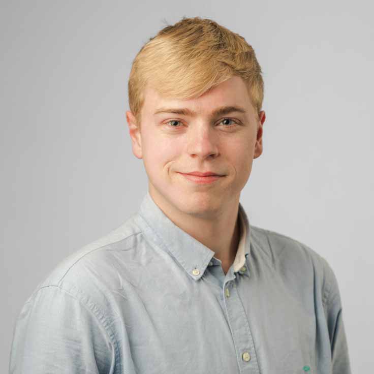 A young man with short blonde hair, wearing a light blue button-up shirt, against a plain grey background.