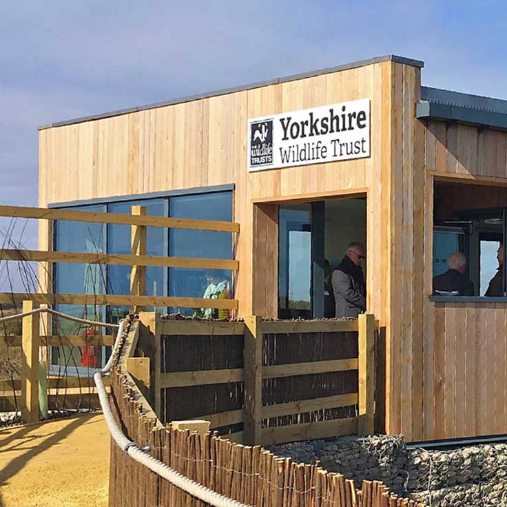 A wooden building of Yorkshire Wildlife Trust with large windows and a sign. Visitors are seen near the entrance.