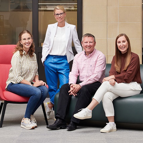 A group of four individuals sits in a modern office setting. They are wearing various professional outfits.