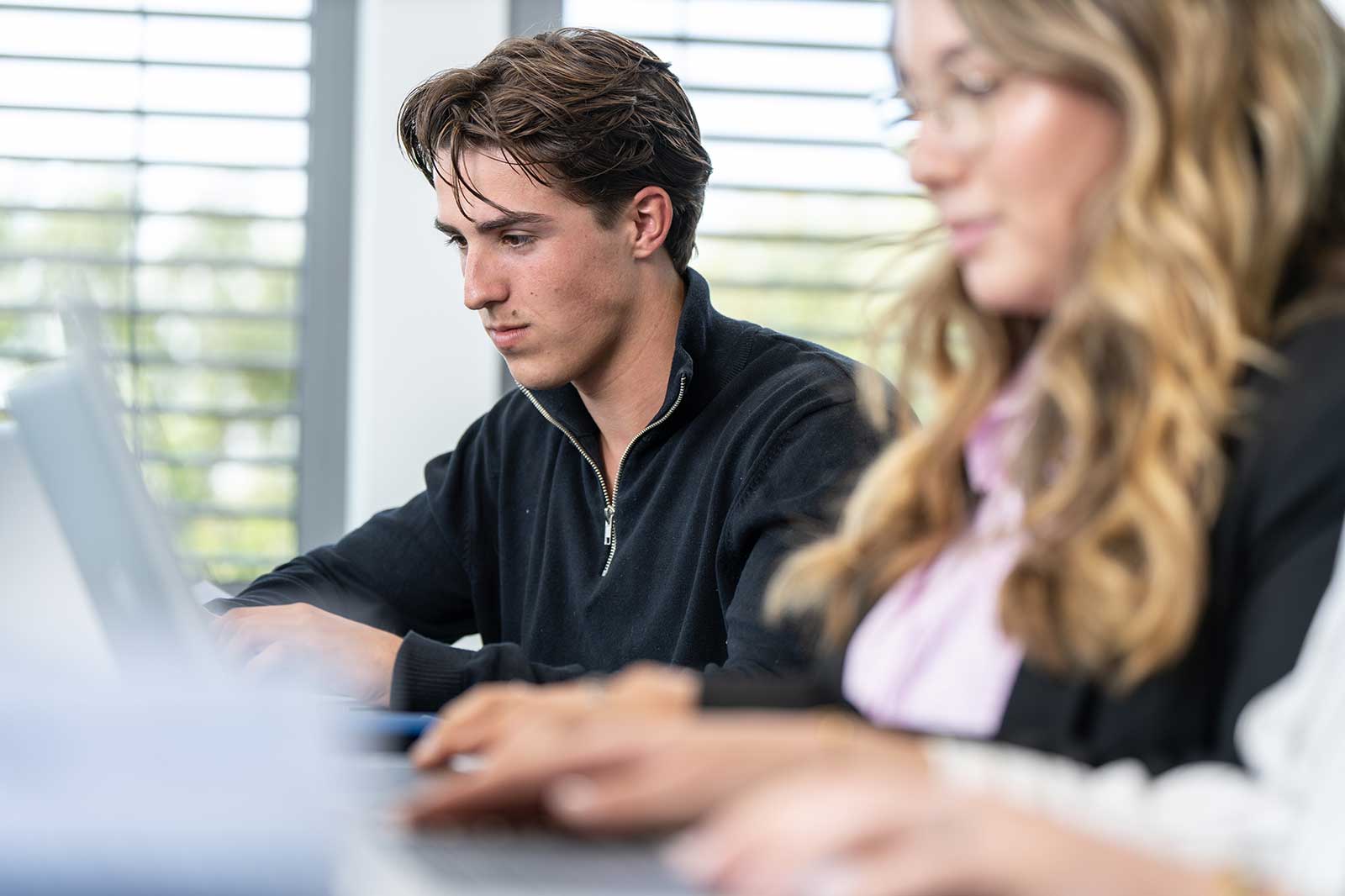 A close-up of two individuals working on laptops in a modern office environment, with natural light filtering through blinds.