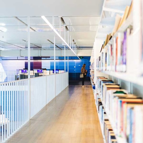 A modern library corridor lined with bookshelves, featuring a person browsing and computer stations in the background.