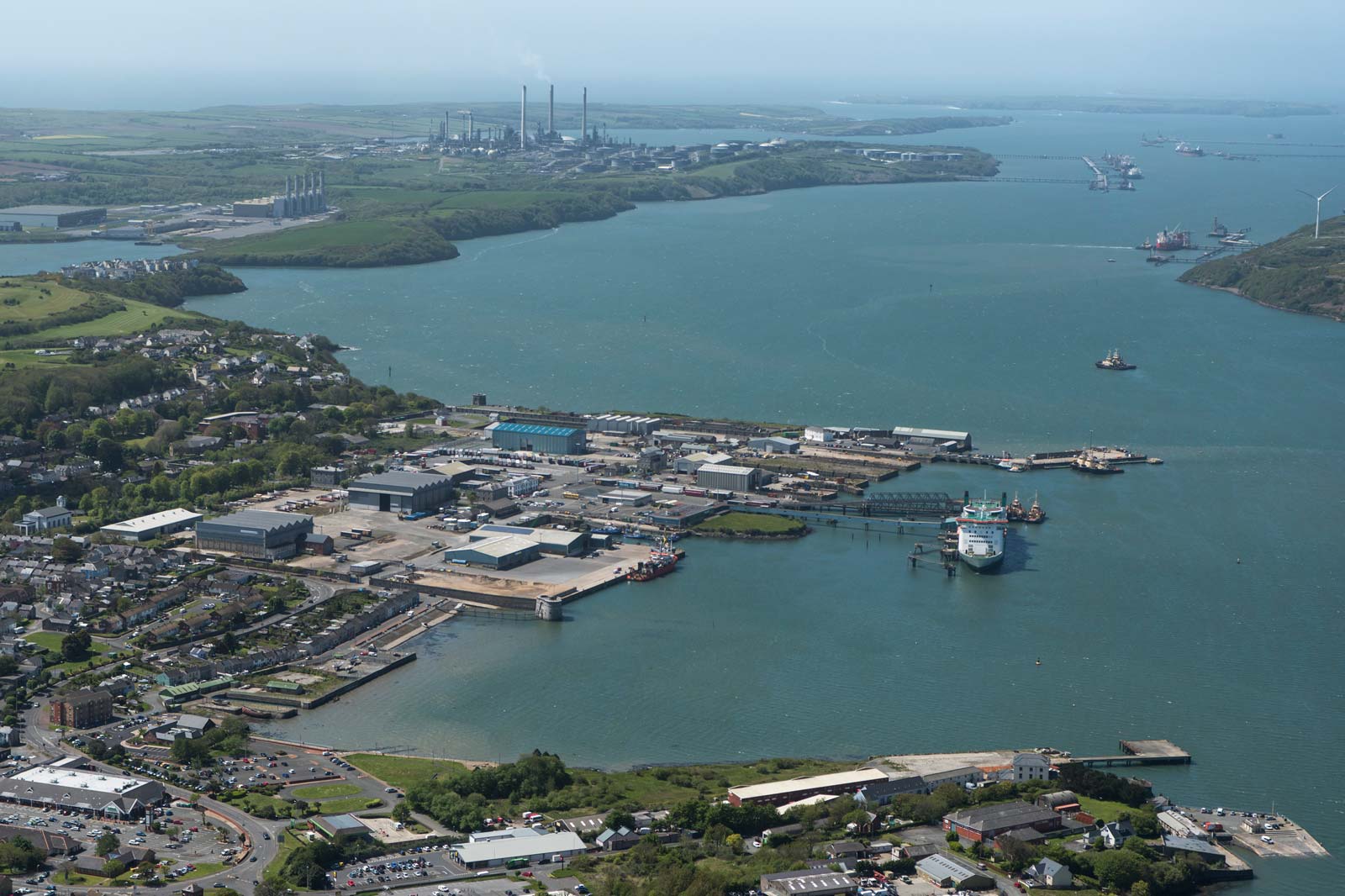 Aerial view of a bustling port with ships, industrial buildings, and green hills under a clear blue sky.