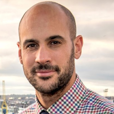 A person with a shaved head and visible ears wears a red, blue and white checkered shirt against a cloudy sky and distant harbor cranes.