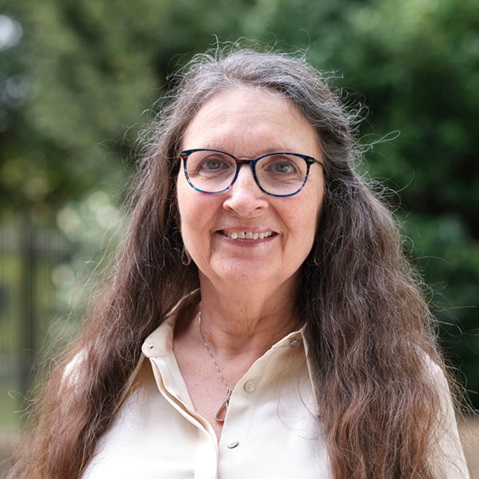 A person with long wavy brown hair wears light cream sleeveless blouse and silver necklace with small pendant, standing outdoors against leafy green foliage. Portrait of Celia Anderson.