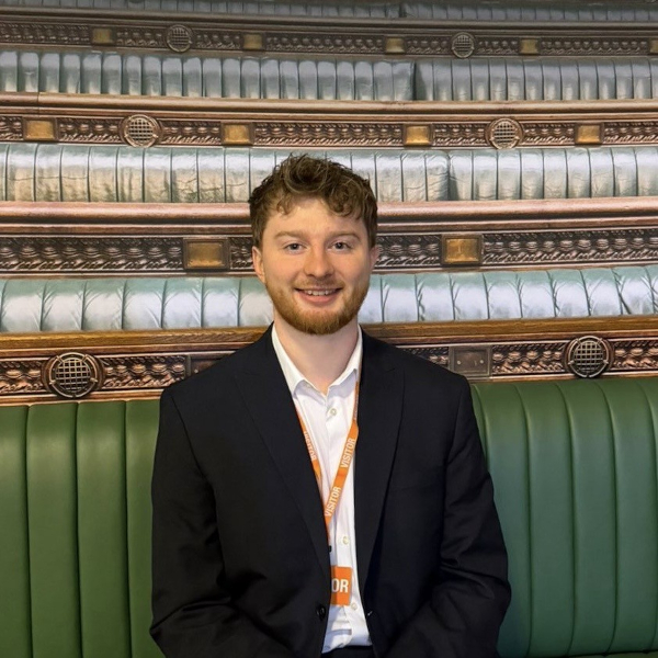A man in a dark suit and white shirt sits on a green bench wearing an orange lanyard labeled "VISITOR" against a decorative wooden backdrop.