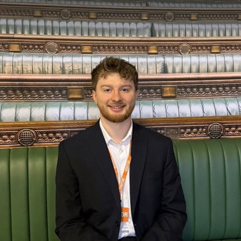 A man in a dark suit and white shirt sits on a green bench wearing an orange lanyard labeled "VISITOR" against a decorative wooden backdrop.