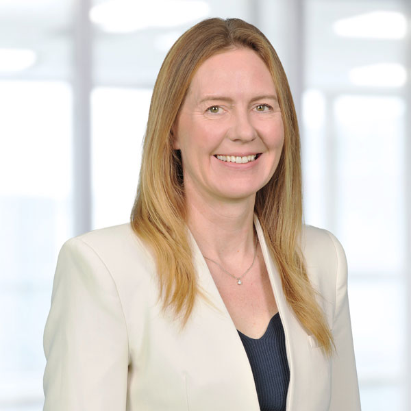 A person with shoulder-length blonde hair wearing a cream blazer, ribbed navy top and small pendant necklace stands against a bright windowed office background.