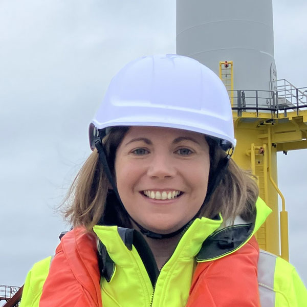 Worker wearing a white hard hat, neon yellow jacket and orange life vest stands before a gray turbine tower and yellow platform under cloudy sky. Portrait of Helen Elphick.