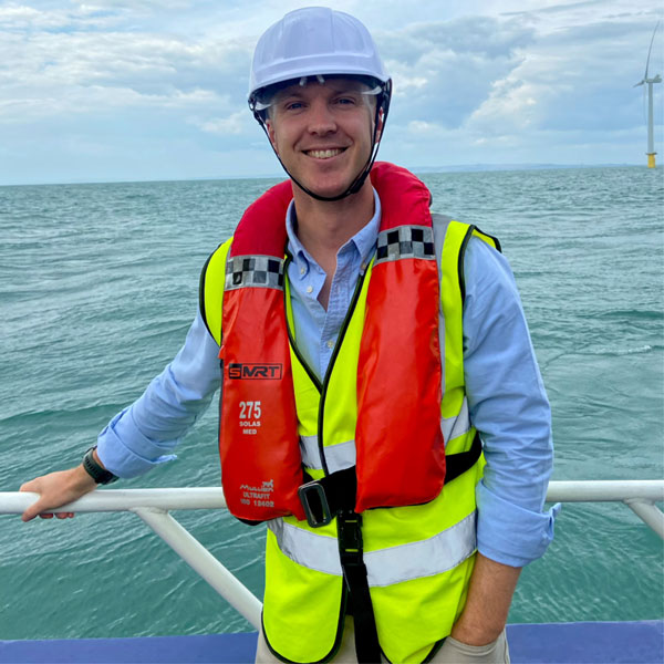 Person wearing a white hard hat, yellow reflective vest and red life jacket labeled "275" leans on boat railing with gray sea and wind turbine. Portrait of James Brabben.