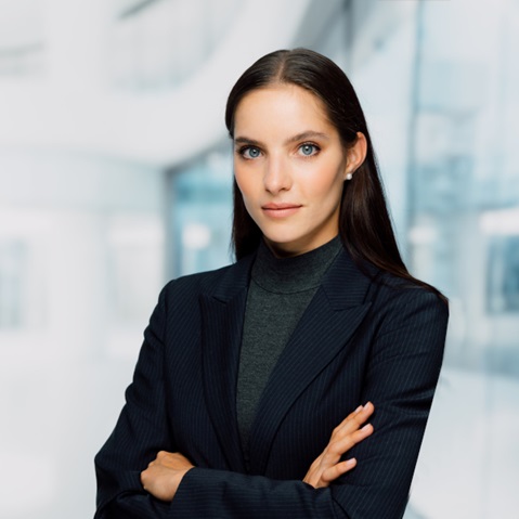 A person with long dark hair stands with arms crossed wearing a pinstripe blazer, charcoal turtleneck and pearl earrings against a bright glass office background.