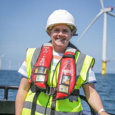 Worker in a white hard hat, neon yellow vest and red lifejacket labeled "WIN-TECH" stands on a boat with wind turbines offshore. Portrait of Nicola Crawford-Percival.