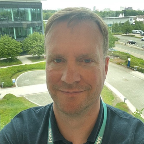 Person in a dark green polo and a green lanyard reading "RWE" stands before a window overlooking an office courtyard with lawns and parked cars.