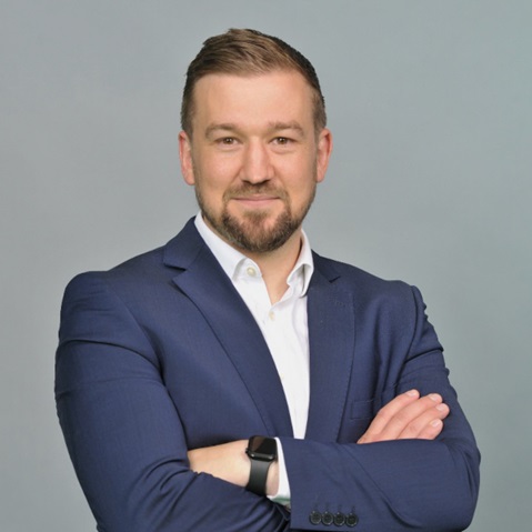 A person stands against a plain light gray background wearing a navy suit jacket and white shirt, arms crossed in a confident professional pose.