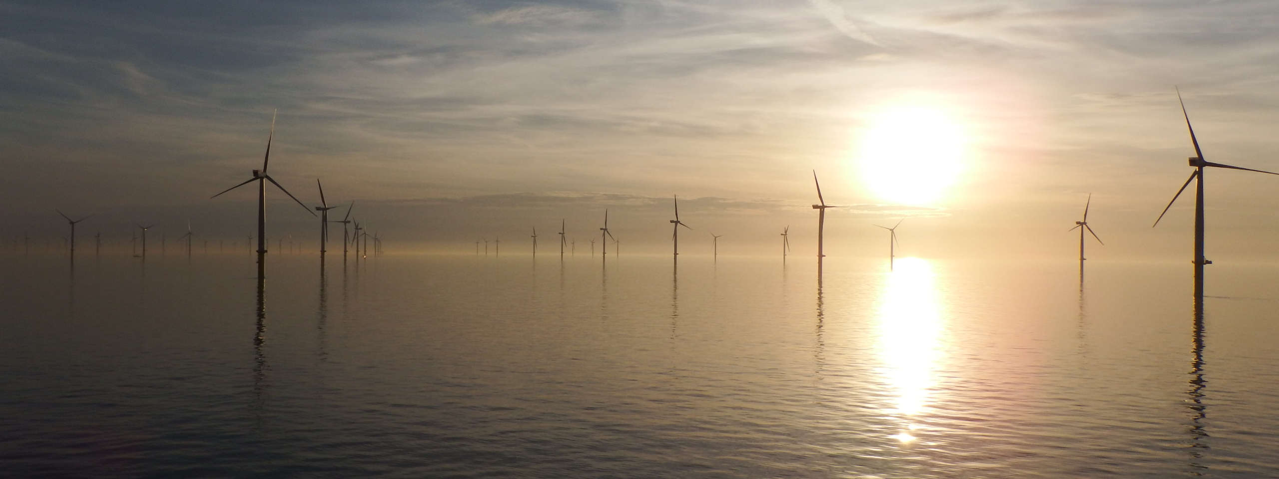 A serene seascape showing multiple wind turbines at sunset, reflected in calm waters with a hazy sky.