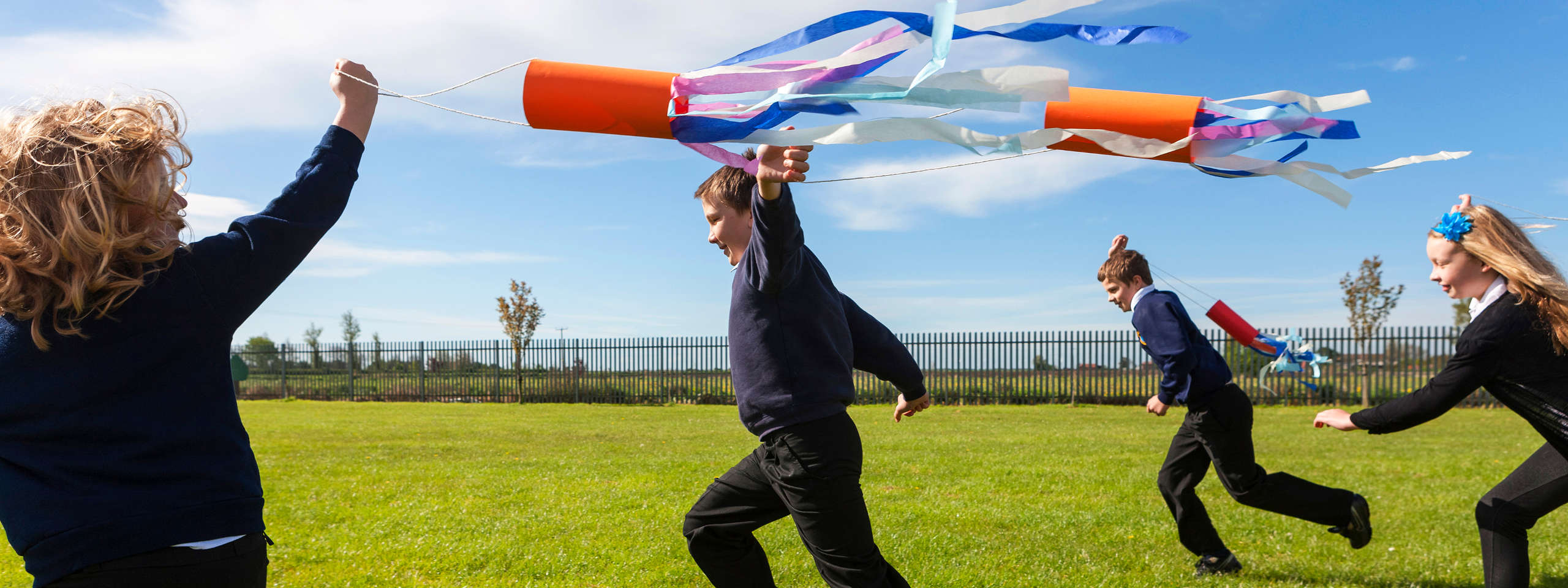 Children play outdoors, joyfully running with a colourful streamer attached to a tube-like object against a blue sky.