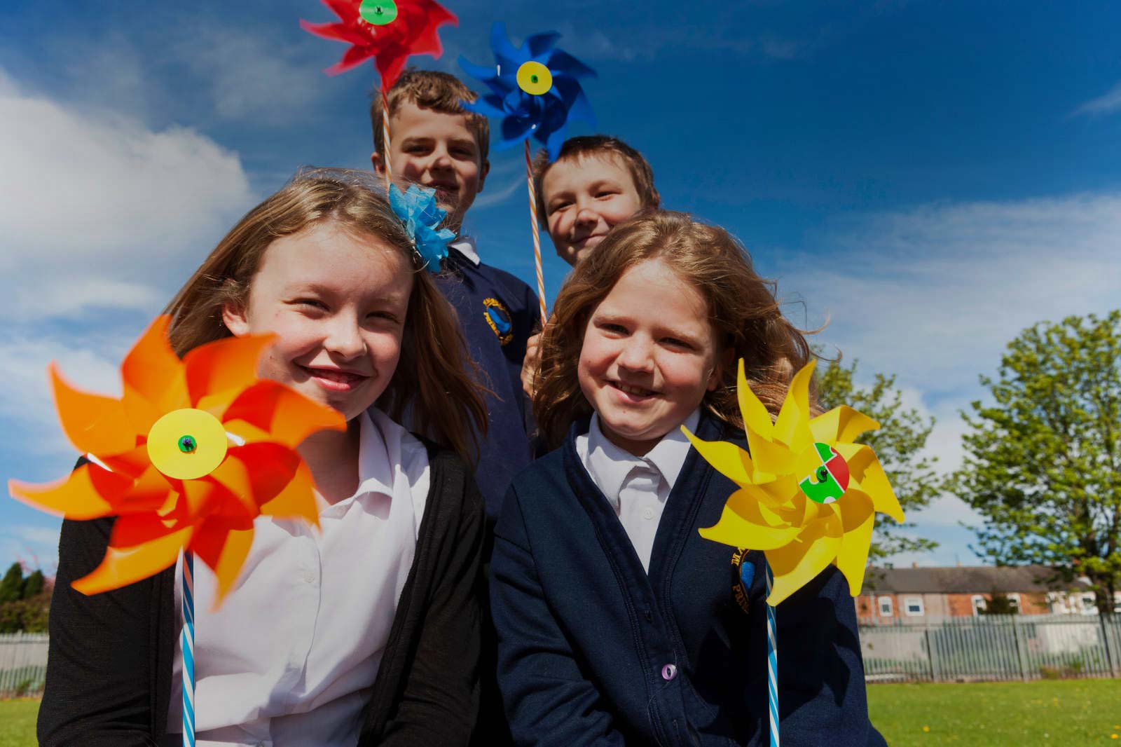 Children in a park hold colourful pinwheels under a bright blue sky, surrounded by greenery and a playground in the background.