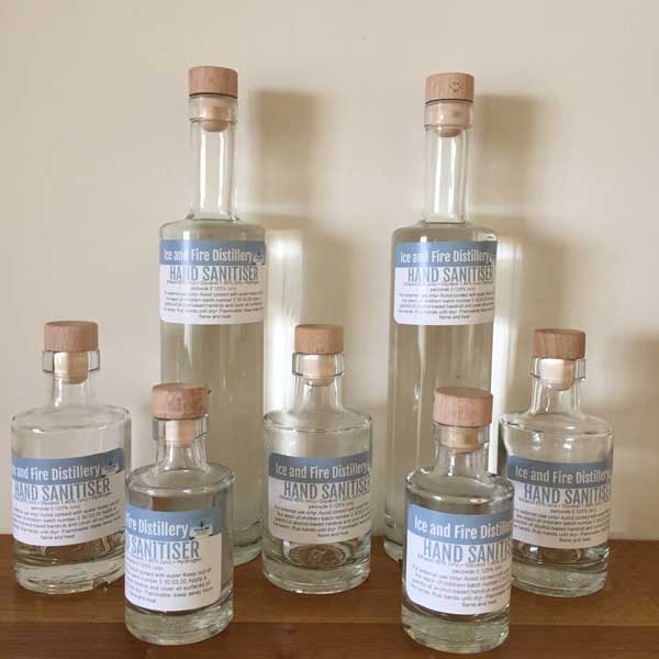 Seven glass bottles of hand sanitiser from Ice and Fire Distillery displayed on a wooden surface.