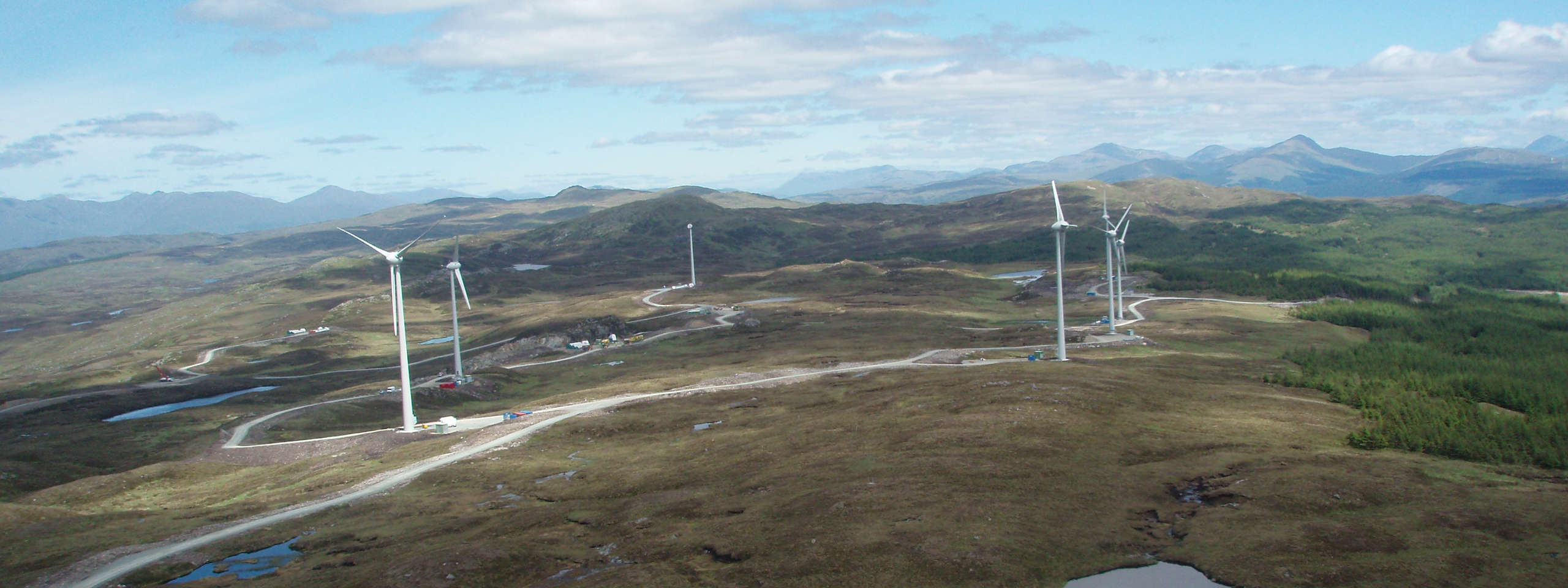 Aerial view of wind turbines on a green hill landscape with mountains in the background and a clear blue sky.