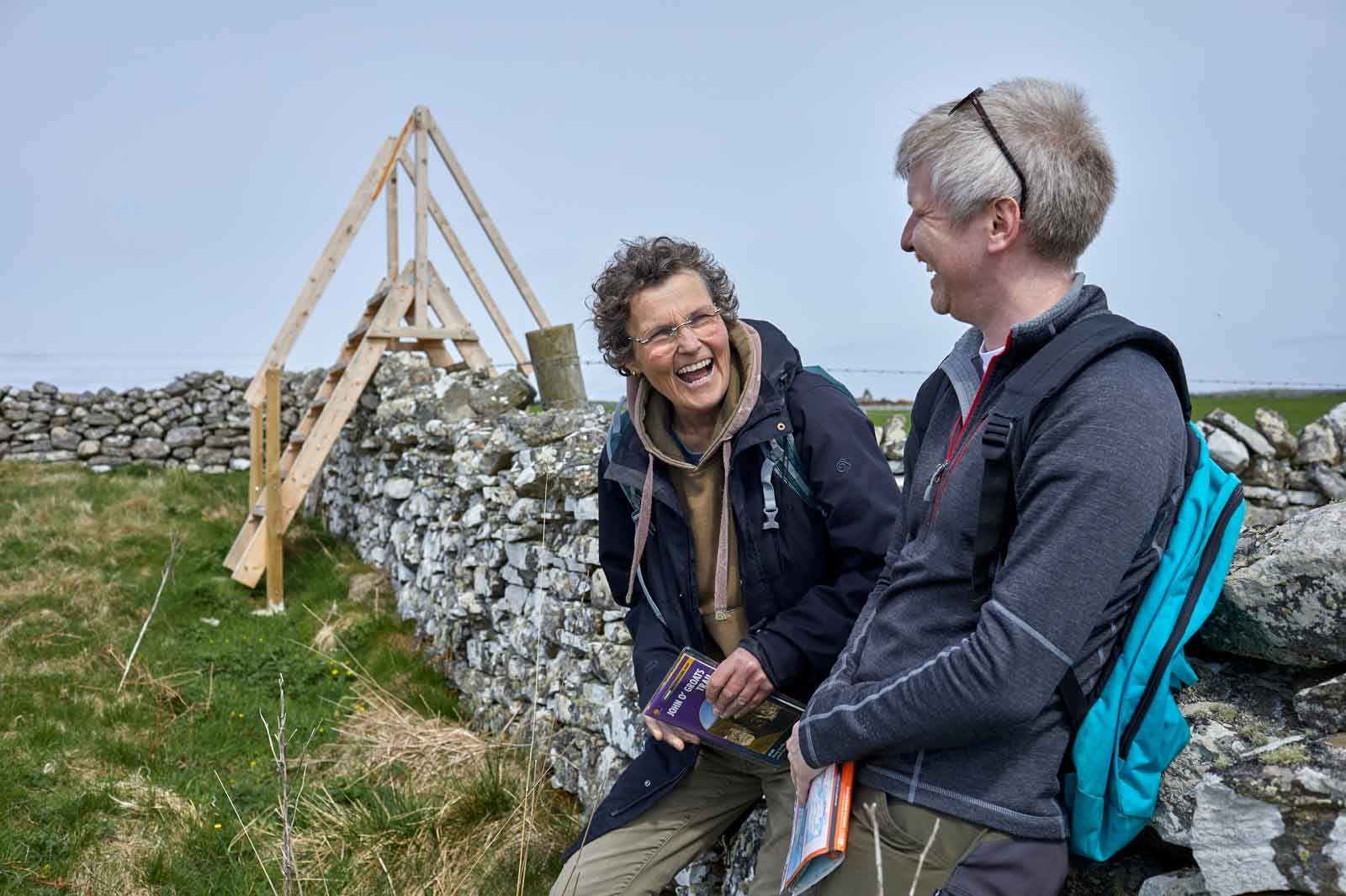 Two people stand near a stone wall, talking while holding guidebooks. A wooden stile is visible in the background.