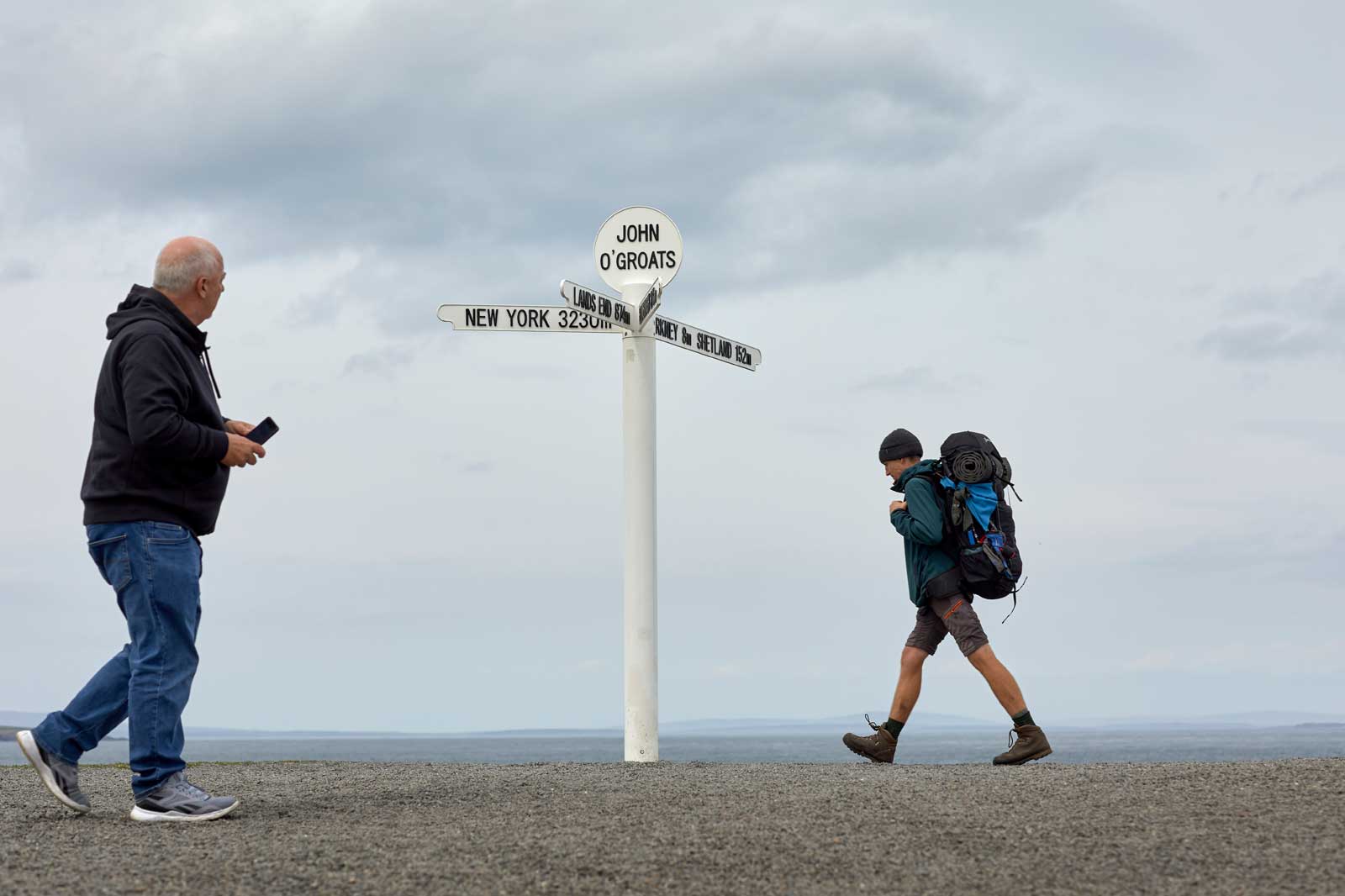 A man walks past a signpost at John O'Groats, showing distances to New York, Lands End, and other locations.