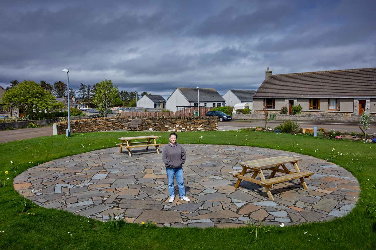 A woman stands in the middle of a circular stone paved area with wooden picnic tables surrounded by grass and houses under a cloudy sky.