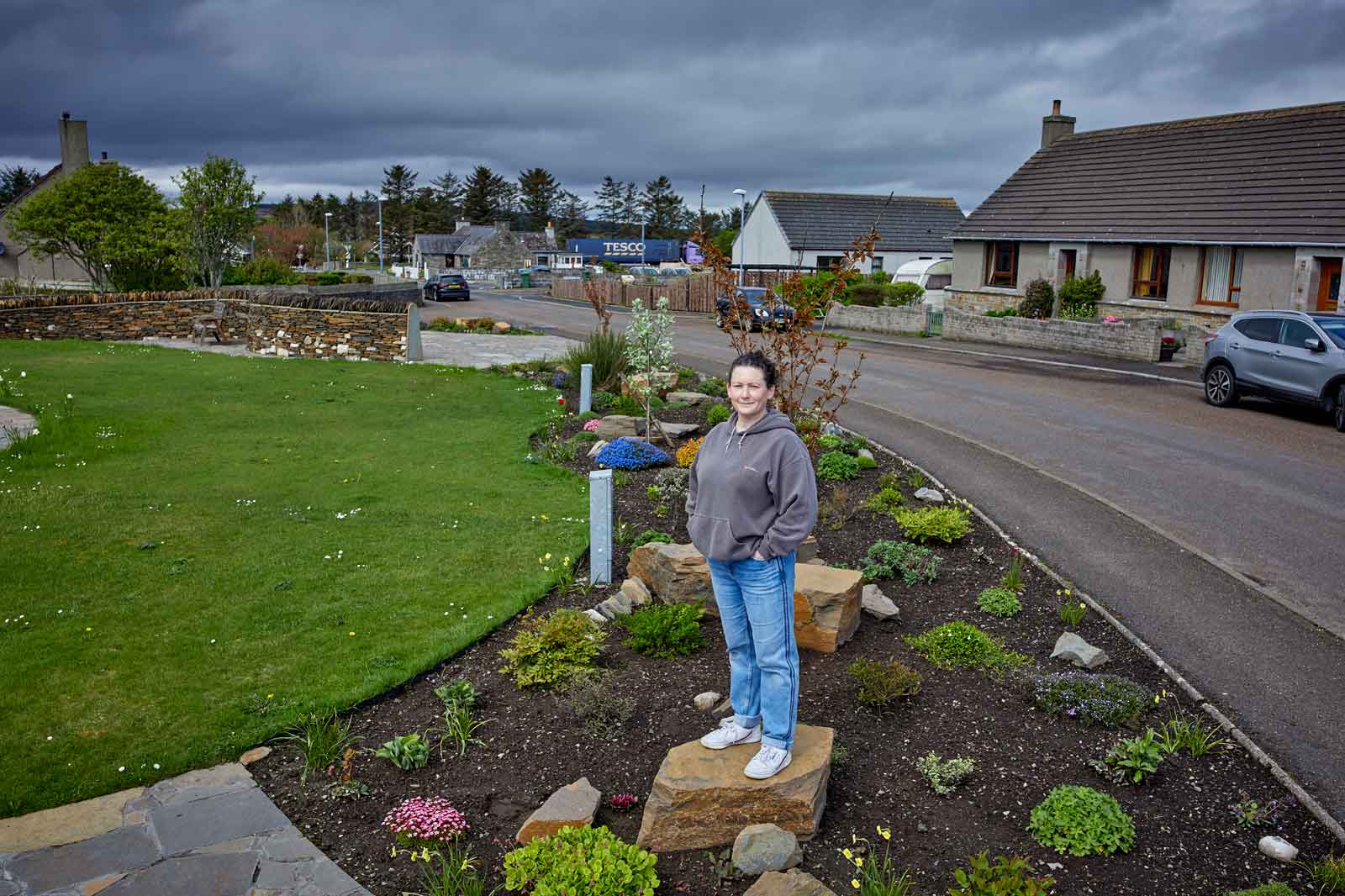 A person stands on a rock in a landscaped garden with flowers, overlooking a road lined with houses under a cloudy sky.