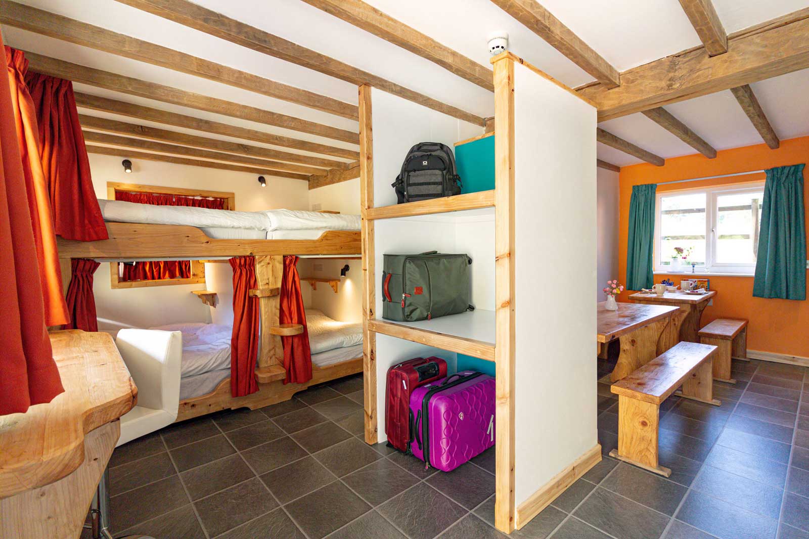 A brightly lit room featuring wooden beams, bunk beds with red curtains, a table, and various bags on shelves.