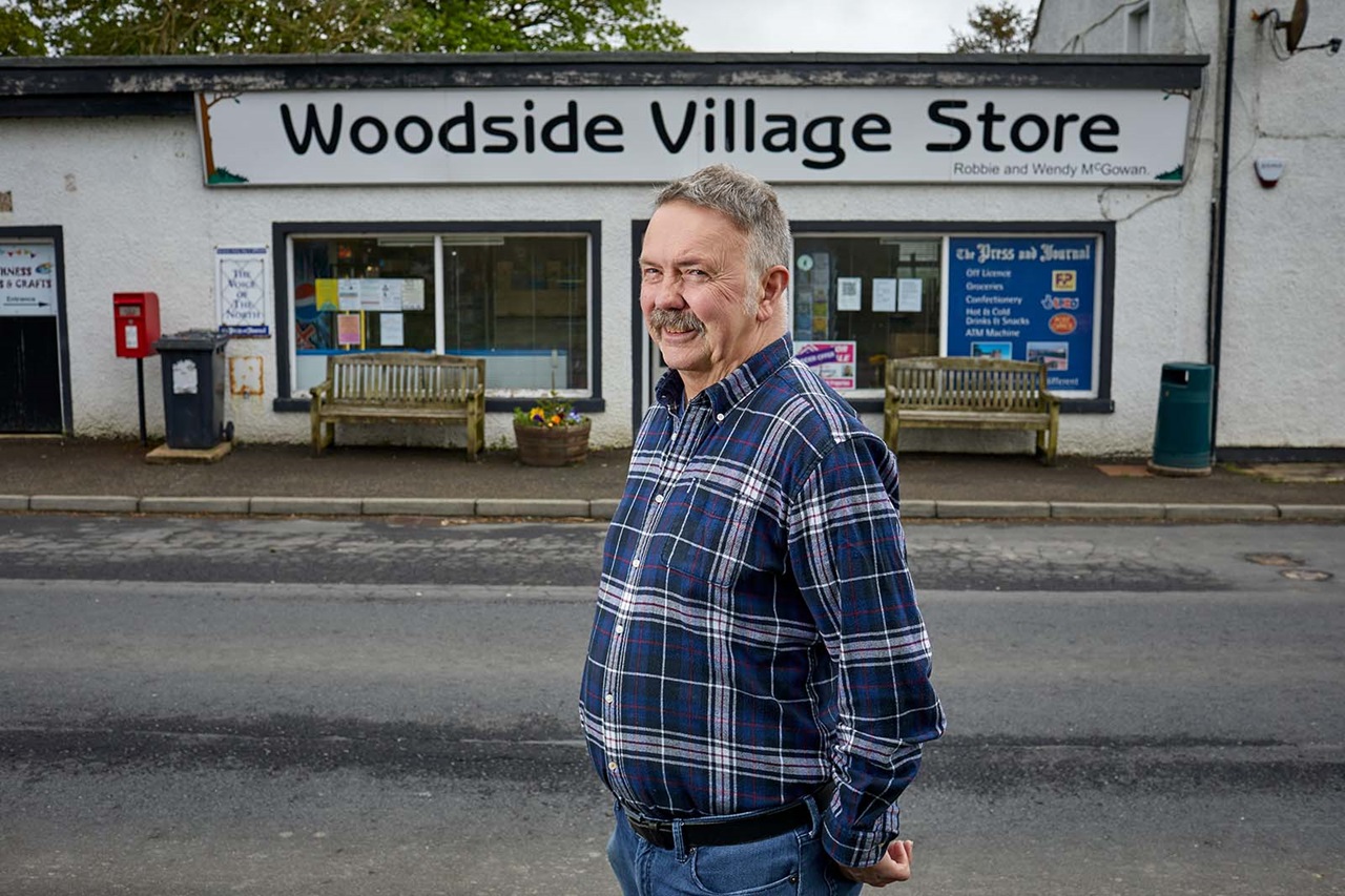 A smiling middle-aged man in a blue plaid shirt stands on the road before a white storefront labeled "Woodside Village Store" with wooden benches.