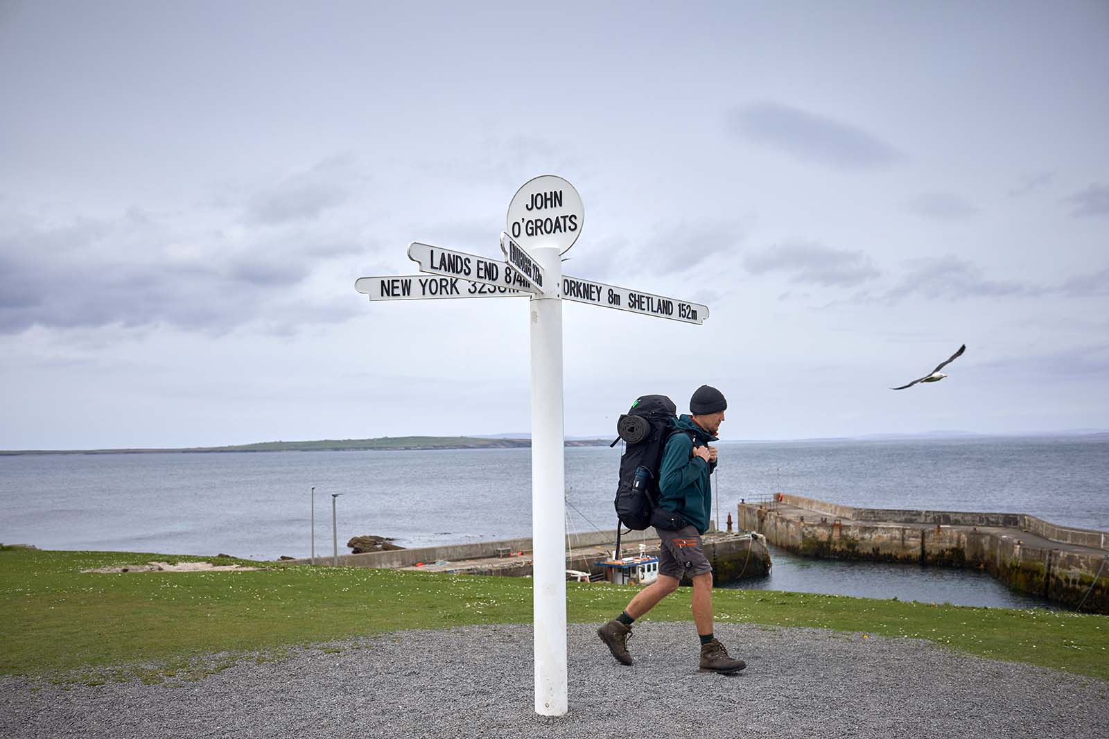A white painted signpost reading "JOHN O'GROATS" stands in the foreground as a backpacker in a teal jacket walks along green grass by a gray sea.
