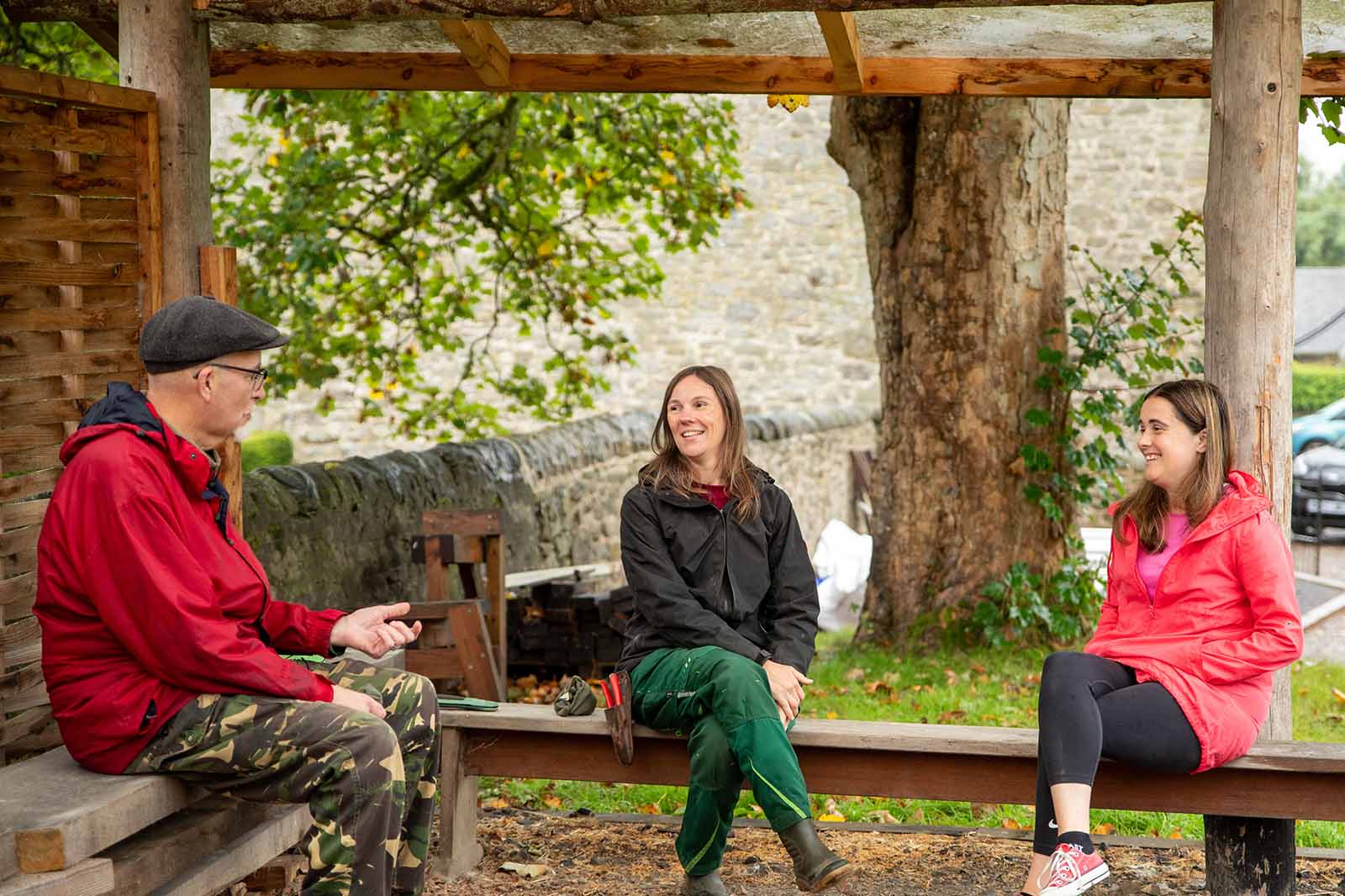 Three people sit on wooden benches under a rustic shelter by a large tree and stone wall, wearing red and black jackets and smiling.