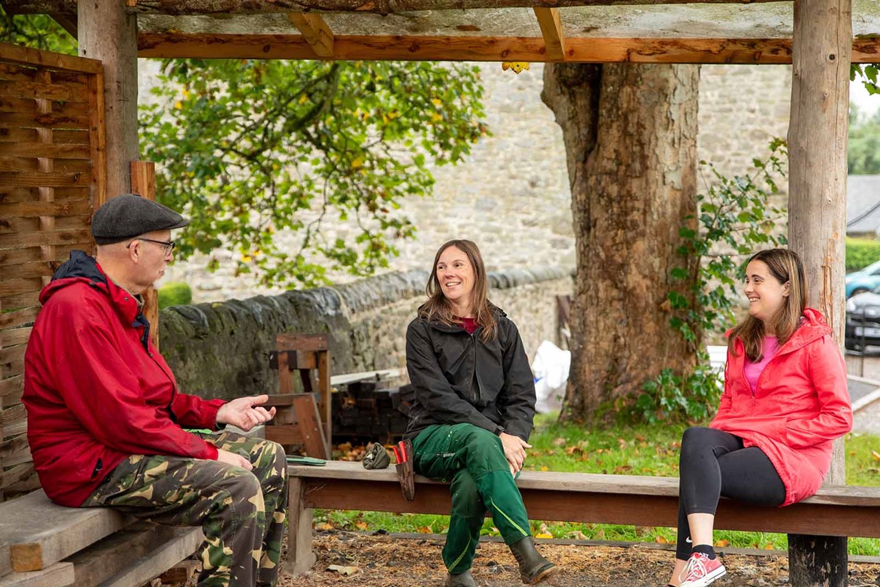 Three people sit on wooden benches under a rustic shelter by a large tree and stone wall, wearing red and black jackets and smiling.