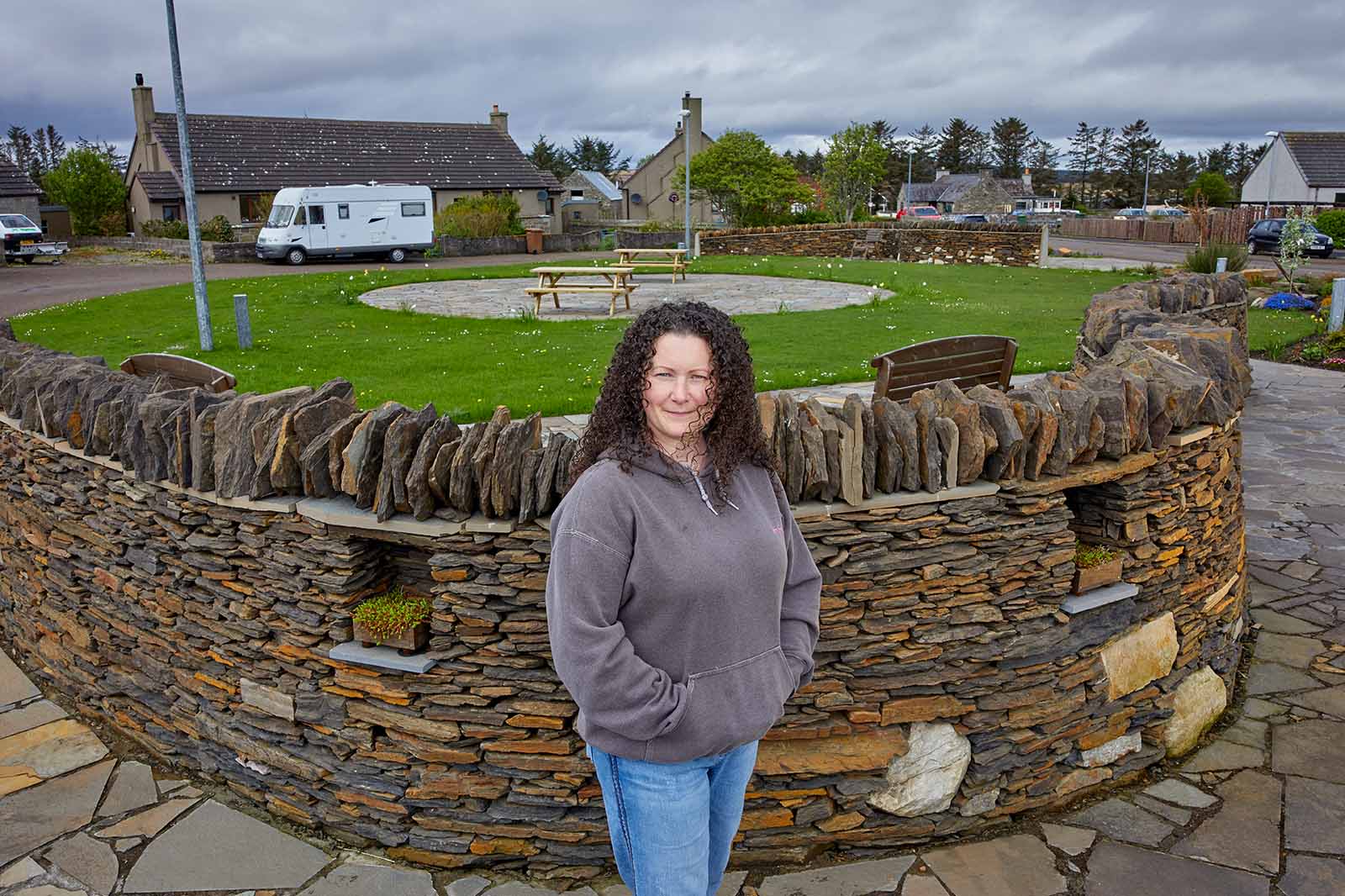 Smiling woman with curly dark hair in a gray hoodie and blue jeans stands by a low stacked-stone wall around a small green park.