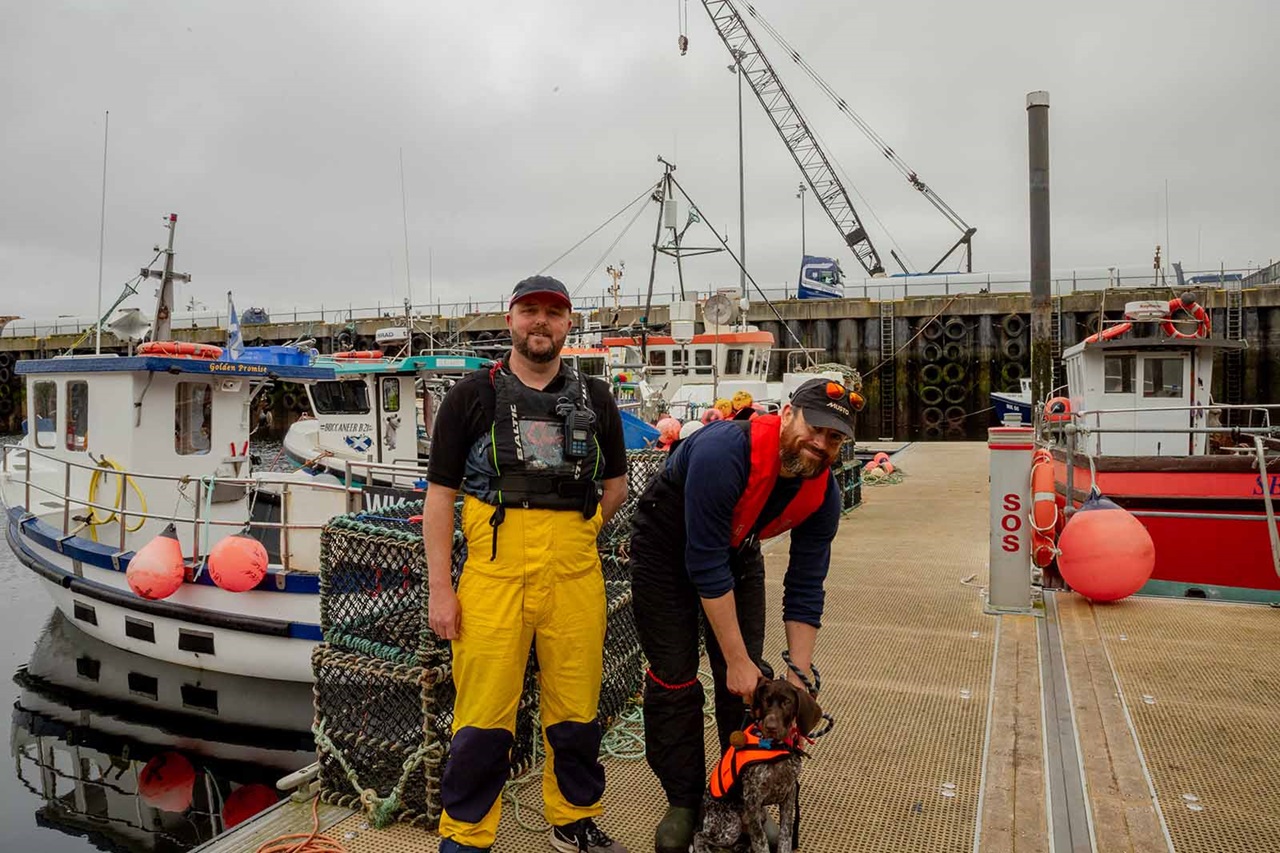 On a dock, two fishermen—one wearing yellow pants—stand by boats and wire crab pots, one bending to pet a dog in an orange life vest.