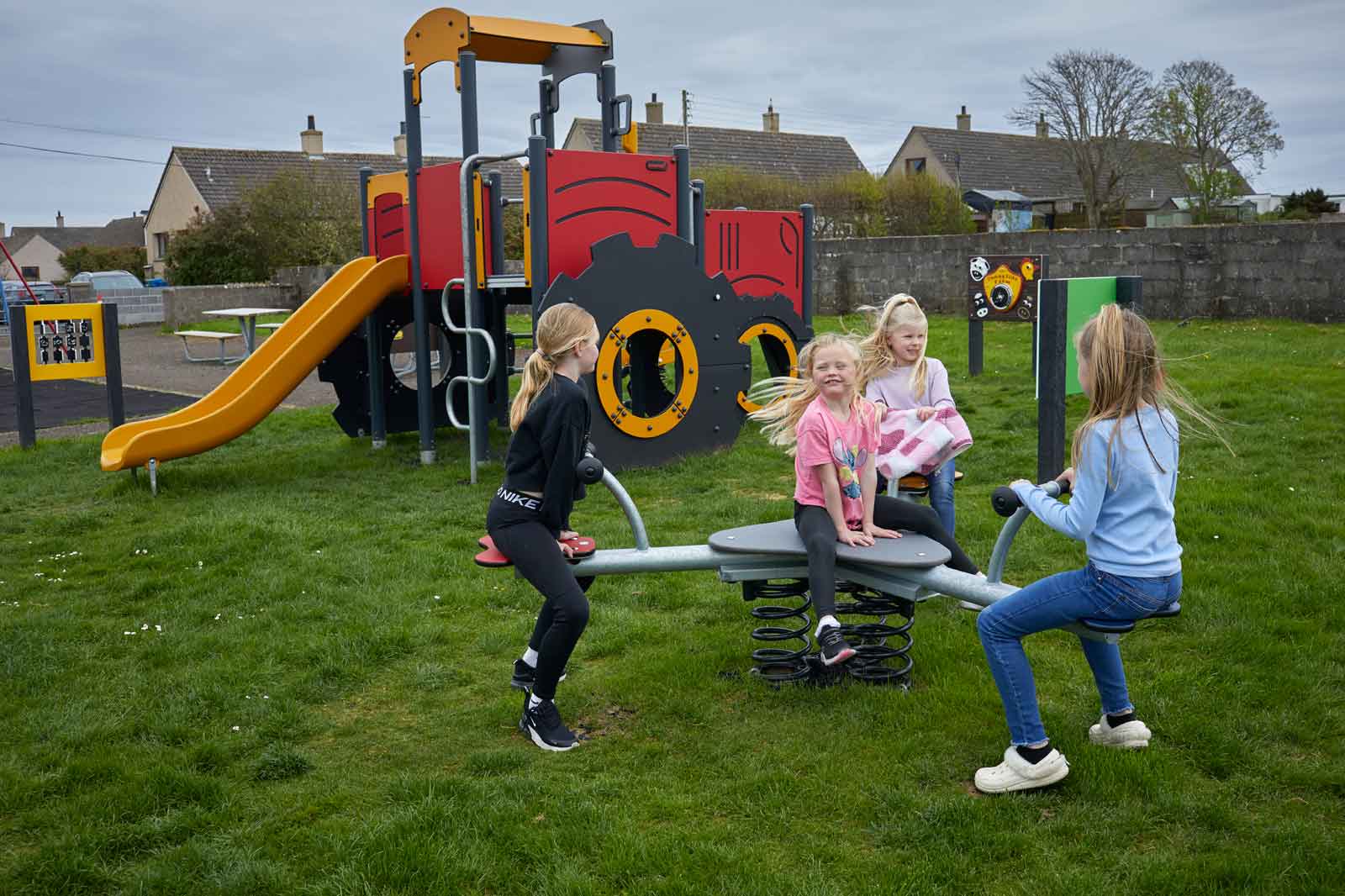 Children play on a playground with a slide, climbing frame, and spring rider in a grassy area with houses in the background.