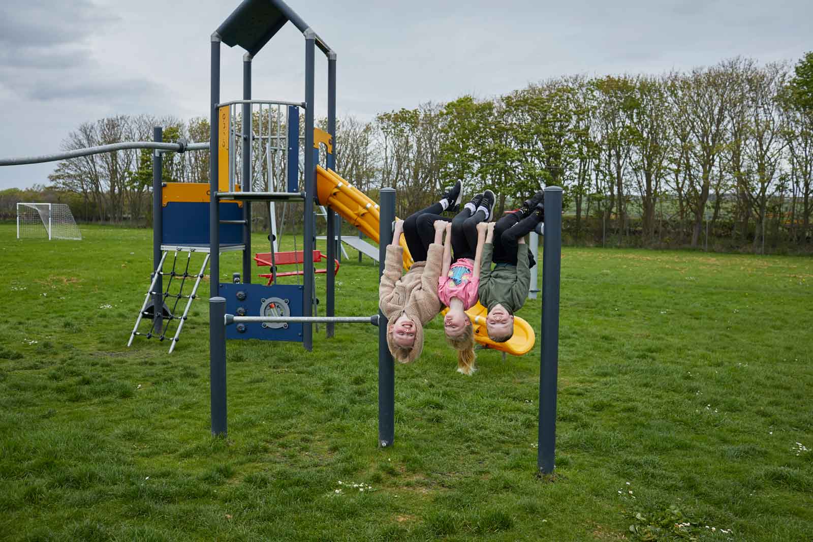 Three children hanging upside down on a playground structure in a grassy field, with trees and a football goal in the background.