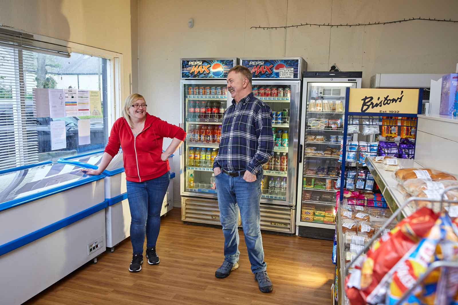 A woman in a red hoodie leans on a counter while a man in a plaid shirt stands nearby in a small shop with coolers and shelves.