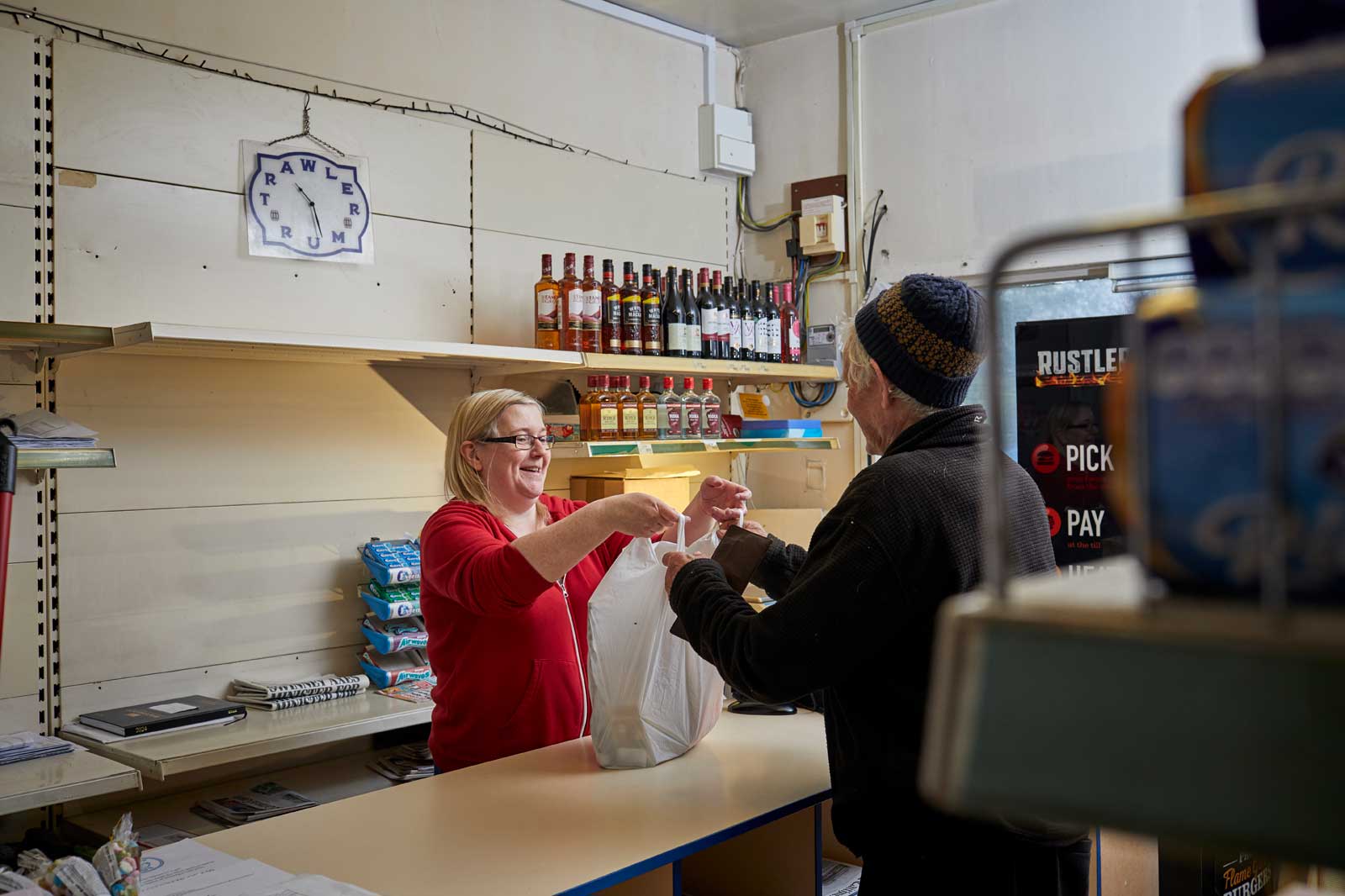 A shop assistant hands a shopping bag to an elderly customer at a counter. Shelves in the background hold various bottles and products.