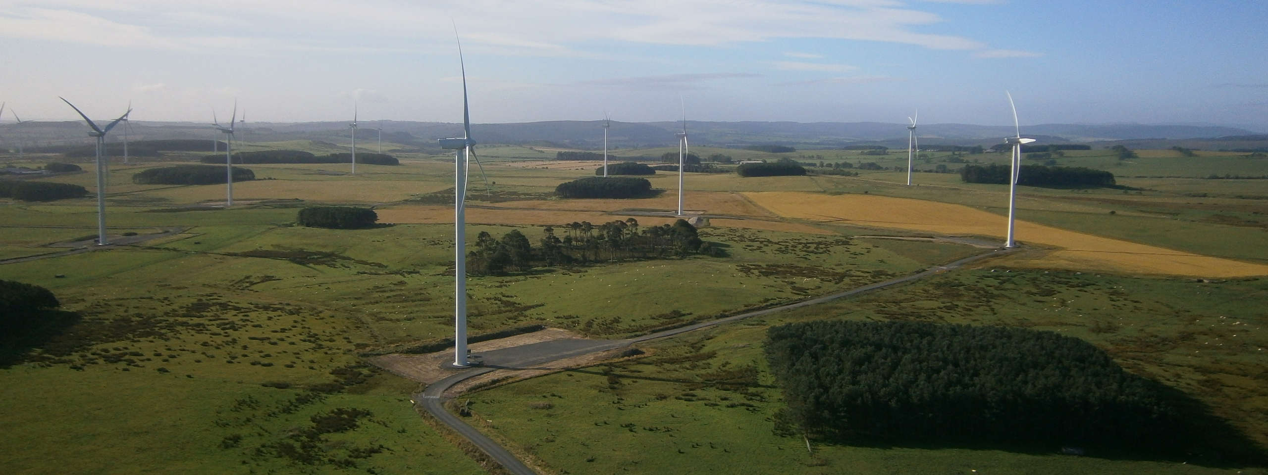Aerial view of a wind farm with multiple wind turbines in a green landscape, surrounded by fields and trees under a clear sky.
