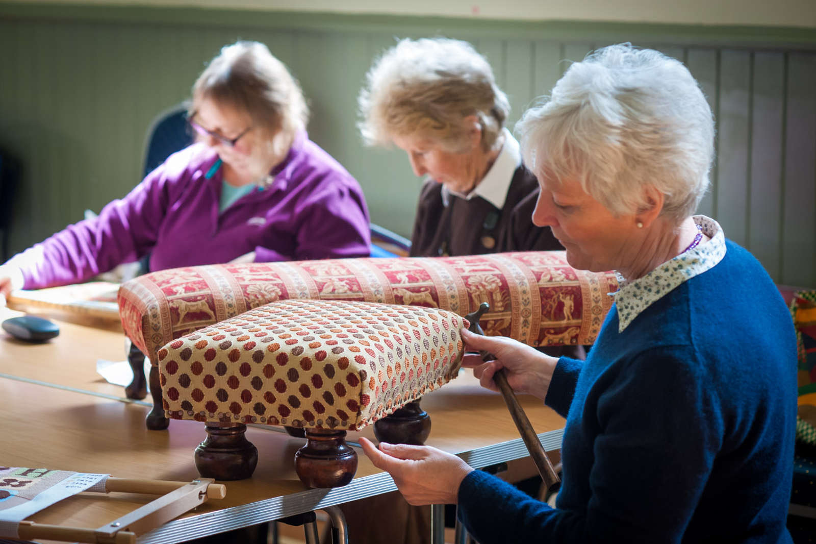A group of women working on upholstered stools at a table in a well-lit room, showcasing various fabric patterns.