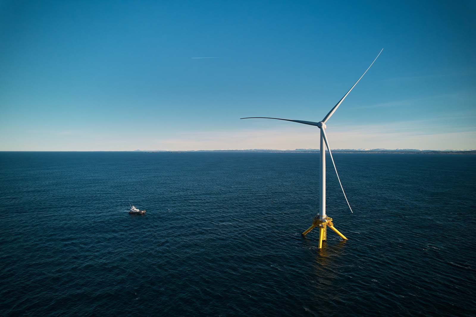 A large wind turbine stands in the ocean with a small boat nearby, under a clear blue sky.