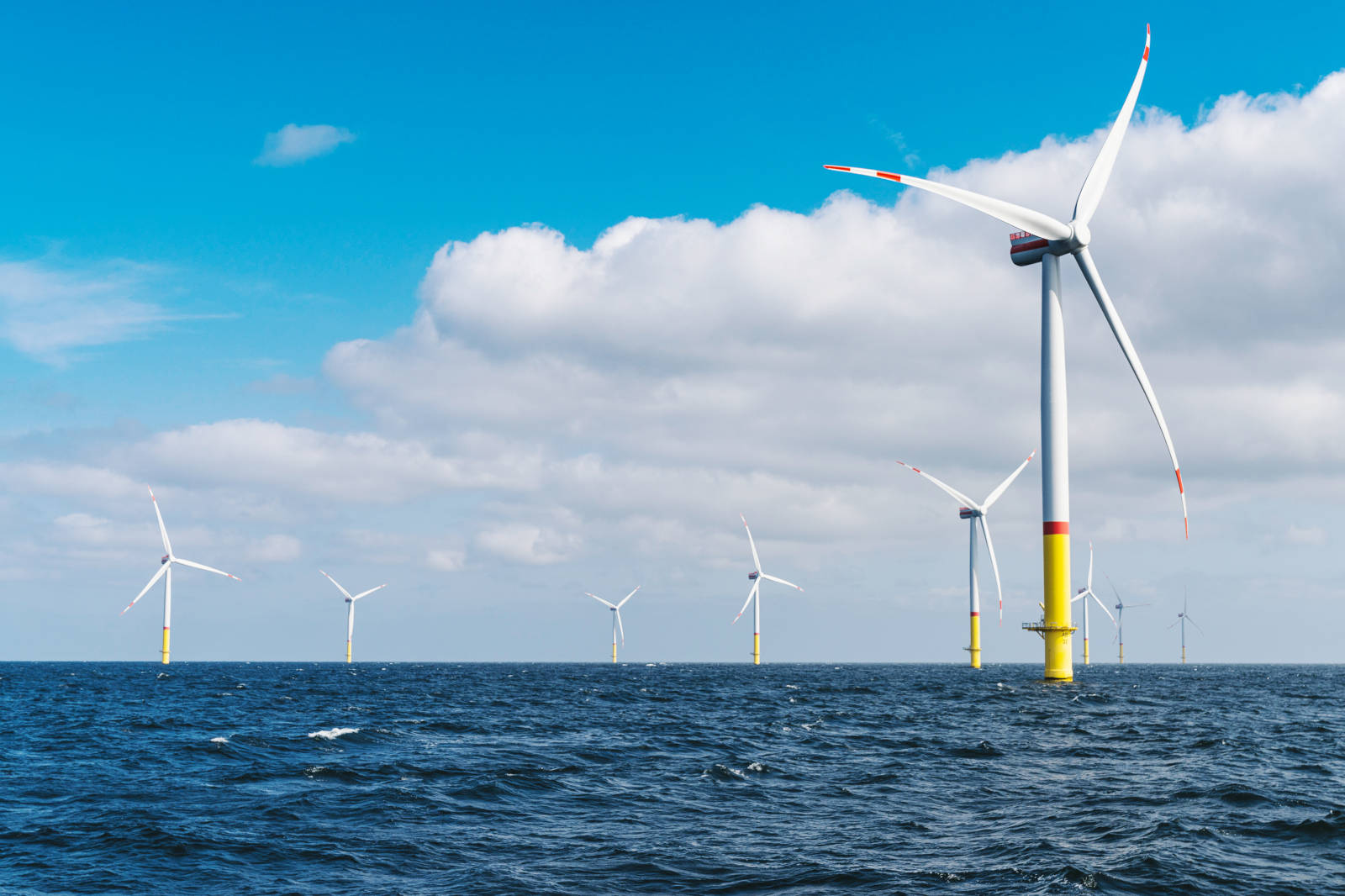 A row of offshore wind turbines in a blue sea under a bright sky with scattered clouds.