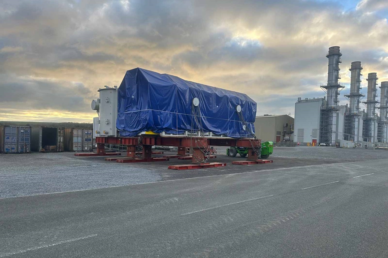A large industrial object covered with a blue tarp, resting on a red platform. The background features a cloudy sky and buildings.