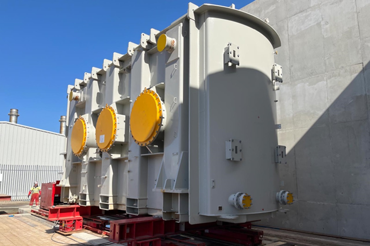 A large industrial transformer with yellow circular valves sits on red rails. In the background, a grey building and blue sky are visible.