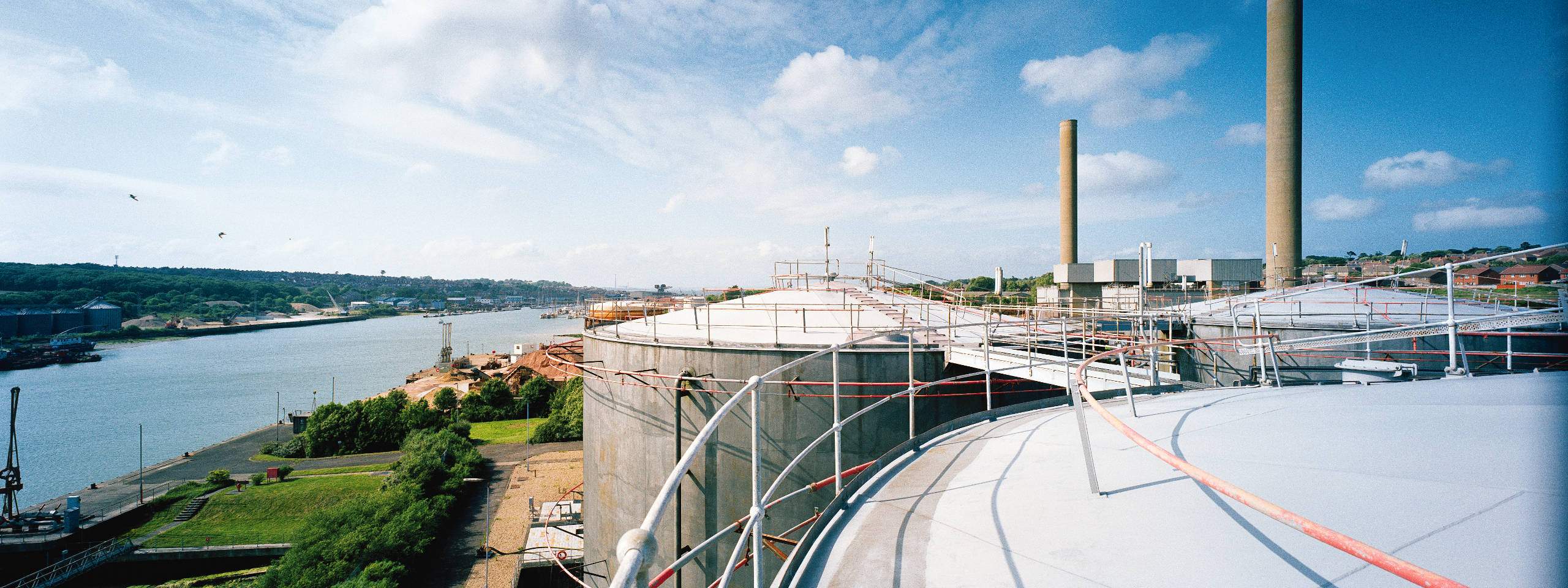 A panoramic view from a facility roof, showing large storage tanks, a river with boats, and distant greenery under a blue sky.